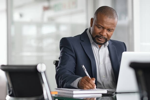 African serious businessman writing notes and using laptop. Mature business man writing his strategy on notebook while using laptop in modern office. Focused black entrepreneur sitting at desk in modern office while working.