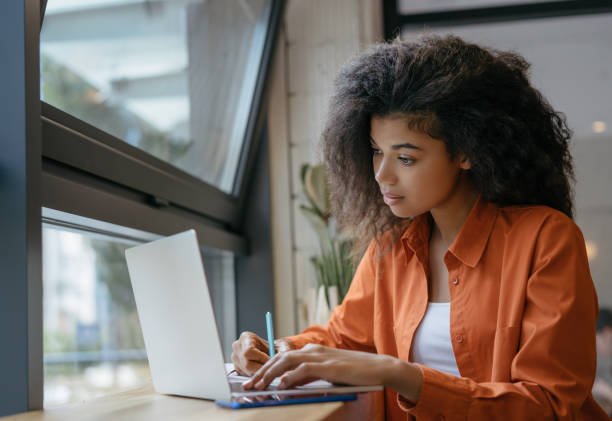 Young student using laptop computer, studying, learning language, exam preparation. Pensive African American woman freelancer writes notes working at home